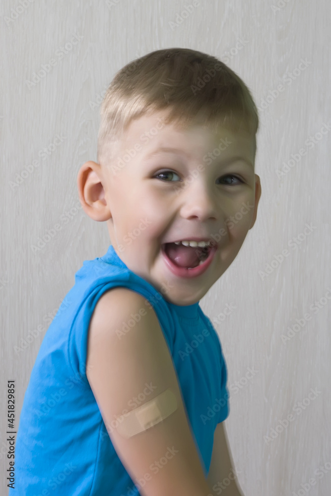 Naklejka premium A cute Boy in a blue T-shirt is laughing merrily, he has a patch on his shoulder, glued after the injection. The vaccine against Covid-19, vaccination of children. Healthcare. Positive emotions