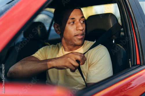 Horizontal shot of the young multiracial man fastening safety belt while sitting and preparing to driving car at the road. Transportation concept photo