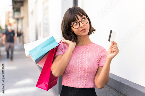 Young brunette woman in the city holding shopping bags and a credit card and thinking