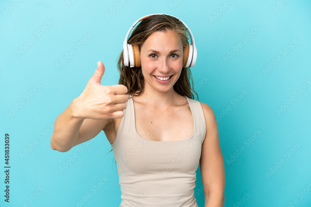 Naklejka premium Young caucasian woman isolated on blue background listening music and with thumb up