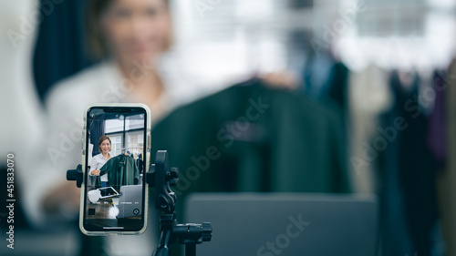 A saleswoman is broadcasting live, selling clothes over the Internet using a smartphone. Select the focus on the smartphone. work at home concept © PIPAT
