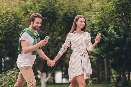 Photo portriat young couple smiling walking in green park holding hands using smartphones © deagreez