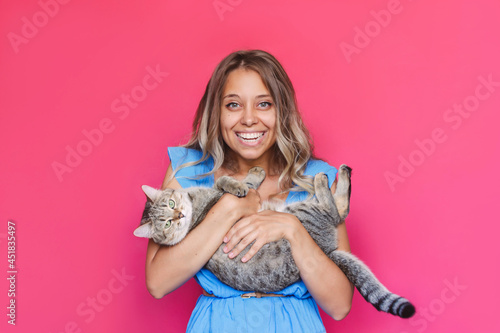 A young caucasian pretty cute blonde woman in a blue dress holds a cat in her hands like a baby isolated on a bright color pink background. The girl babying with a kitten. Fiendship of pet and owner photo