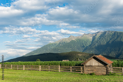 a wooden  cabin near a corn field surrounded by wooden fence with mountains in a background