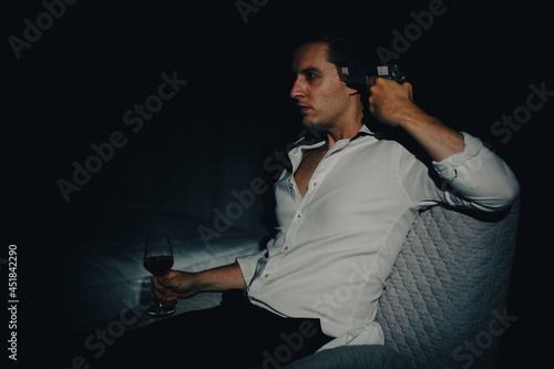 A young man in casual business clothes sitting alone in dark bedroom holding a pistol aim at his head and wine glass looks like going to kill himself. Concept of hopelessness and give up people photo