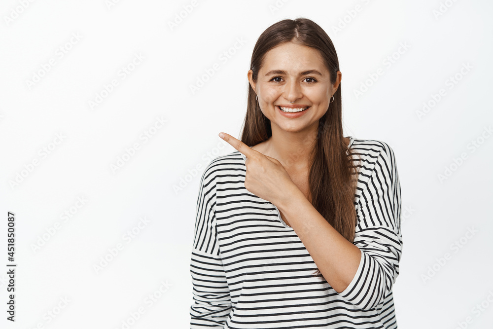 Real people. Young smiling woman pointing finger at upper left corner, showing logo or sale discount info, standing over white background
