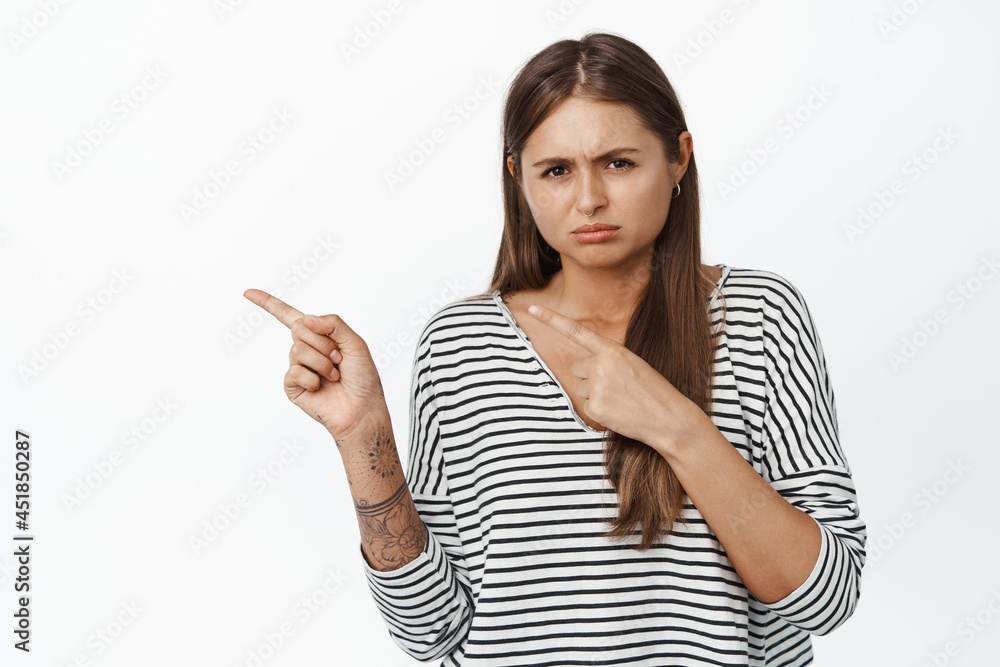 Upset sulking girl pointing fingers left and sobbing, looking disappointed or sad, standing over white background Upset sulking girl pointing fingers left and sobbing, looking disappointed or sad, standing over white background