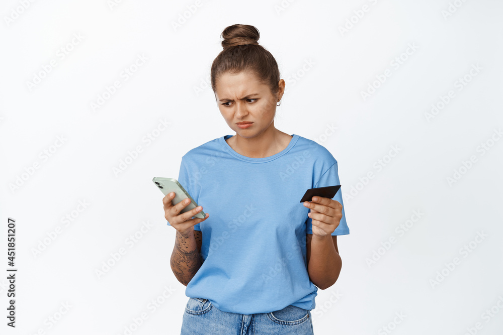 Image of girl looking confused at mobile phone screen and frowning perplexed, holding credit card, problem with online payment, white background Image of girl looking confused at mobile phone screen and frowning perplexed, holding credit card, problem with online payment, white background