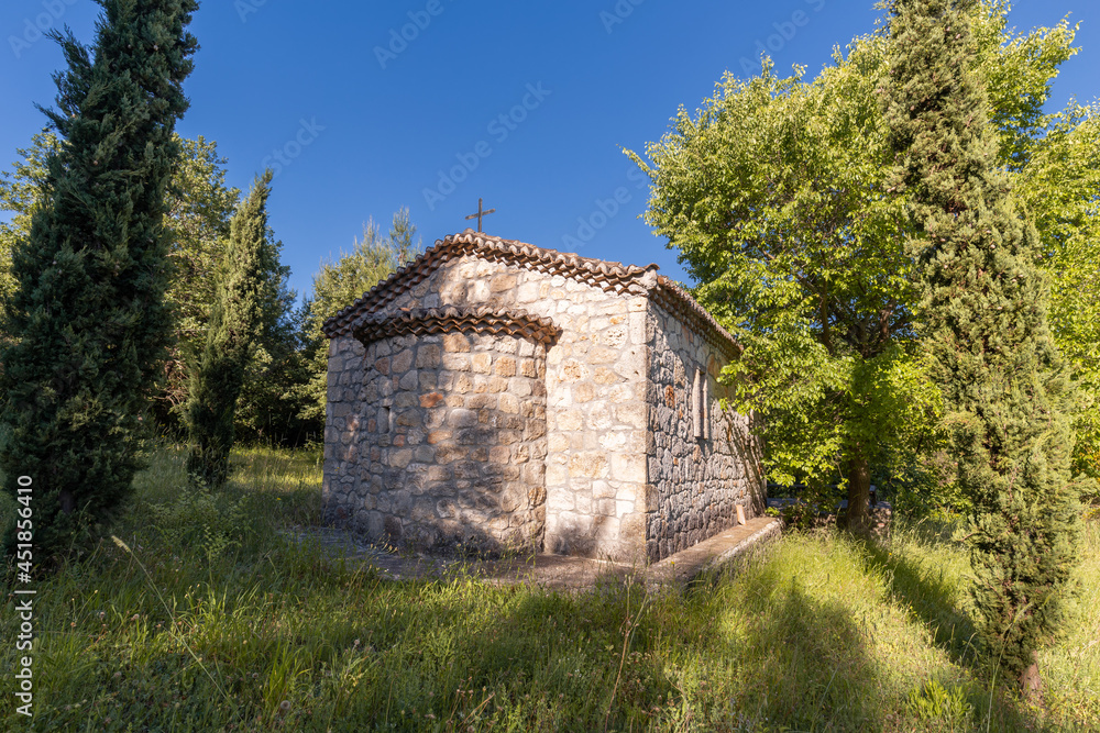 A small old orthodox church with stone walls and a tiled roof in the middle of a tiny meadow surrounded by trees against the clear vivid sky