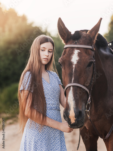 A beautiful young girl with long hair stands next to a horse in nature in the summer
