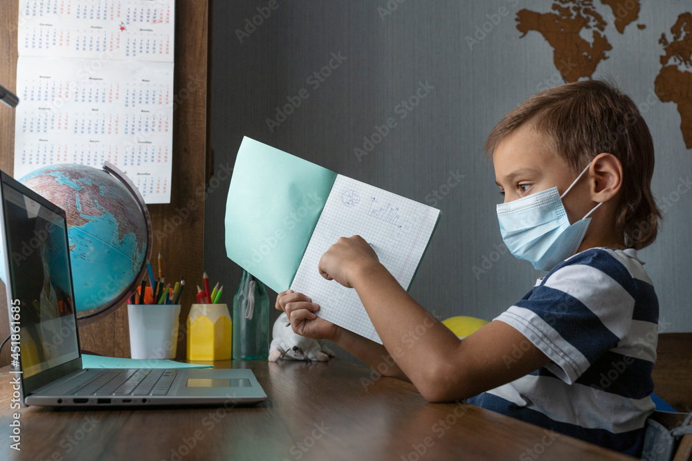 © Anna - Schoolboy during distant online lesson. He is showing homework in notebook to his teacher via laptop screen. Education during pandemic