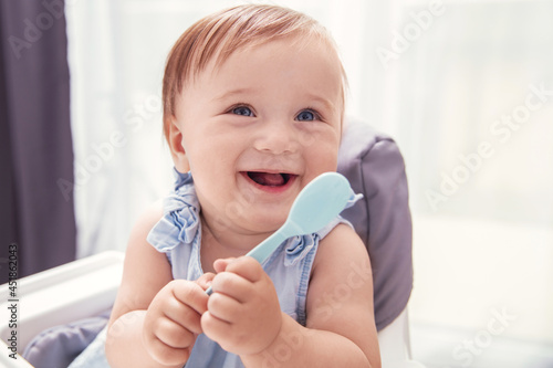 Infant baby girl in blue dress holds baby spoon and smiles photo