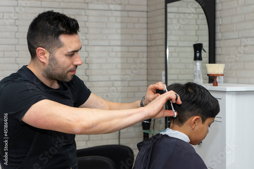  Smiling barber using the scissors to cut the hair of a quiet boy sitting in the barber's chair