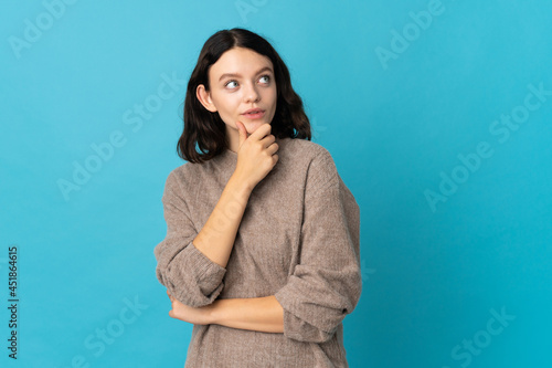 Teenager Ukrainian girl isolated on blue background having doubts