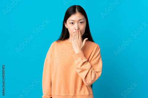 Young Chinese girl over isolated blue background covering mouth with hand