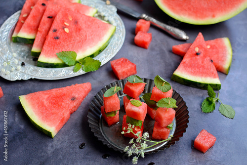 Whole and sliced watermelon on a metal plate and green mint leaves on a dark gray background.