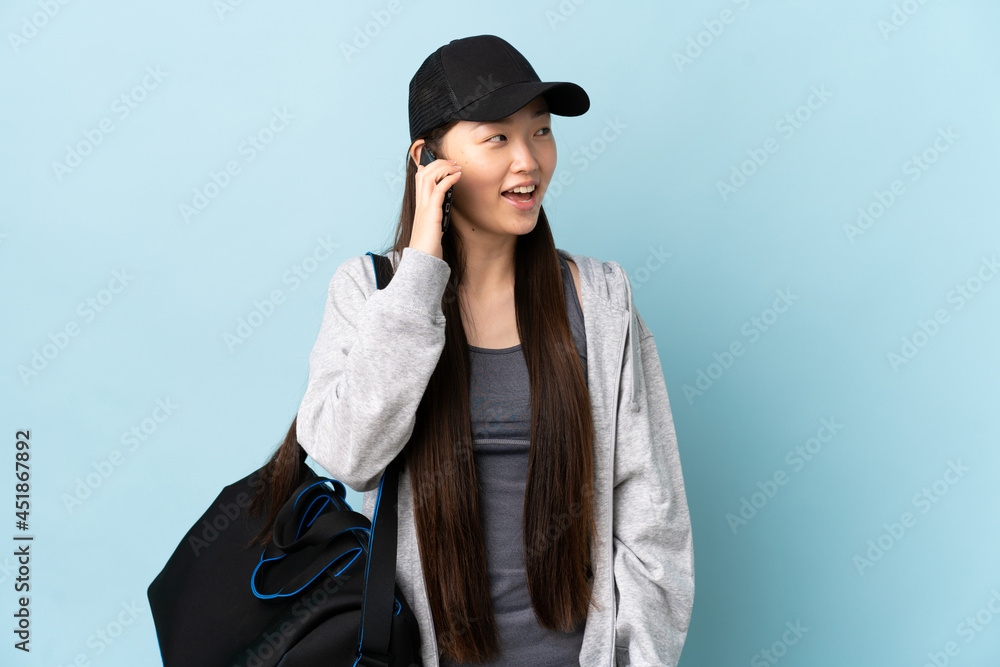 Young sport Chinese woman with sport bag over isolated blue background keeping a conversation with the mobile phone Young sport Chinese woman with sport bag over isolated blue background keeping a conversation with the mobile phone