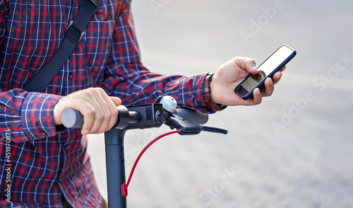 Young man wearing shirt leaning on electric scooter handlebar, holding mobile smartphone in his hands, closeup detail
