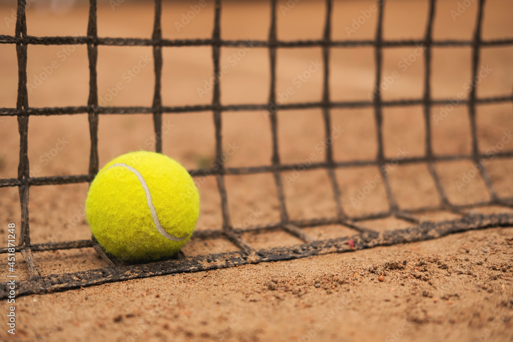 A new tennis ball rests on a net on a sandy court.
