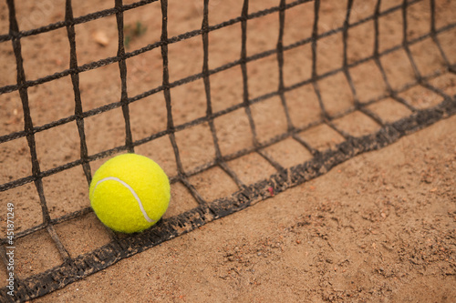 A new tennis ball rests on a net on a sandy court. © Илья Мышенков