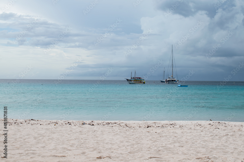 A view from a beach on the west coast of Barbados. There is white sand and blue water with a few boats in the sea.