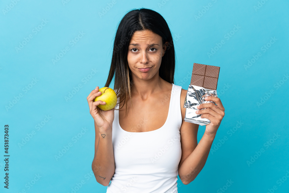 Caucasian girl isolated on blue background taking a chocolate tablet in one hand and an apple in the other Caucasian girl isolated on blue background taking a chocolate tablet in one hand and an apple in the other