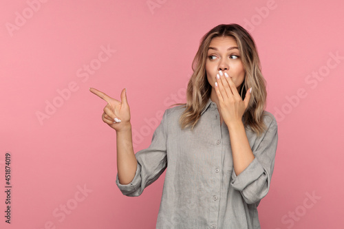 Pretty shocked blonde female pointing finger to empty cope space presenting shocking advice information, sale poster or ads covering open month with hand. Excited young girl isolated over studio wall