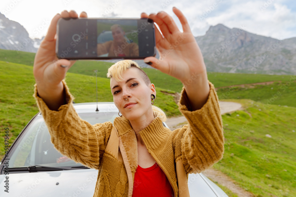Happy young Caucasian girl with punk style and piercings takes selfies during a stop on her car trip up the mountain.