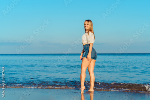 Fitness smiling woman in white top and jeans shorts posing on blue sea and sky background during a walk on the seaside beach. Summer holidays, vacation. Copy space.