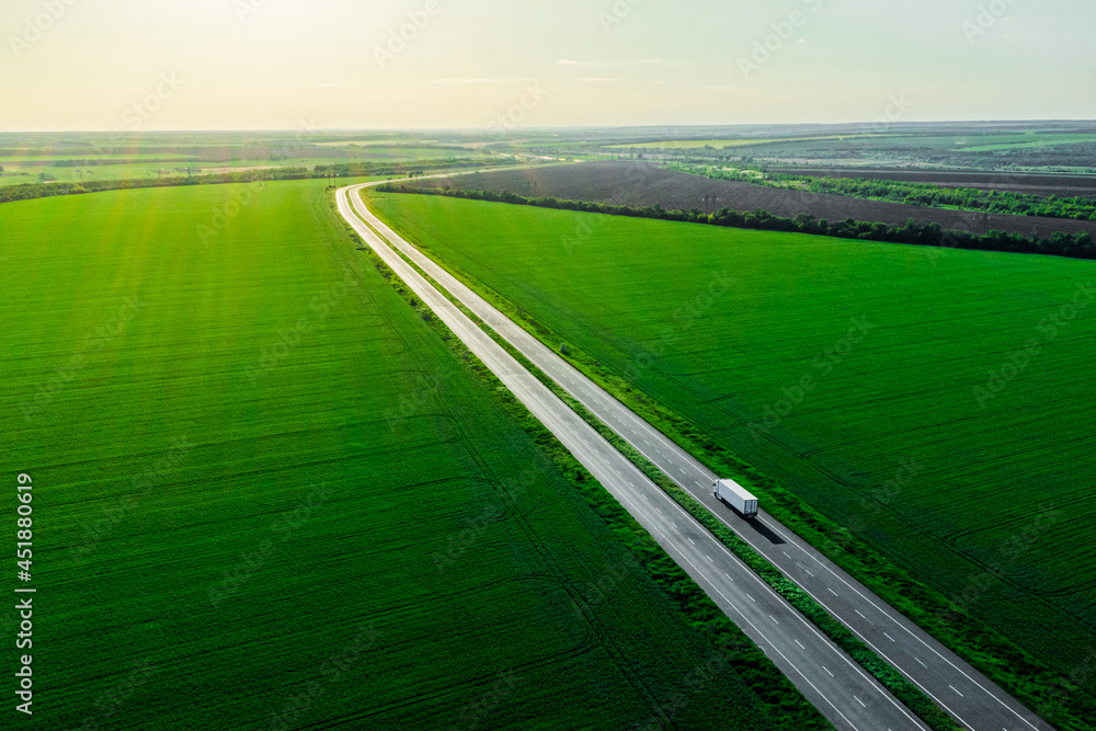 Fototapeta premium alone white truck on the higthway at sunset. cargo delivery driving on asphalt road among the green fields with goods. seen from the air. Aerial view landscape. drone photography.
