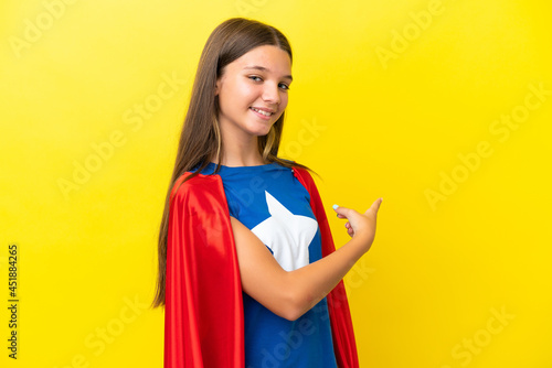Little caucasian superhero girl isolated on yellow background pointing back