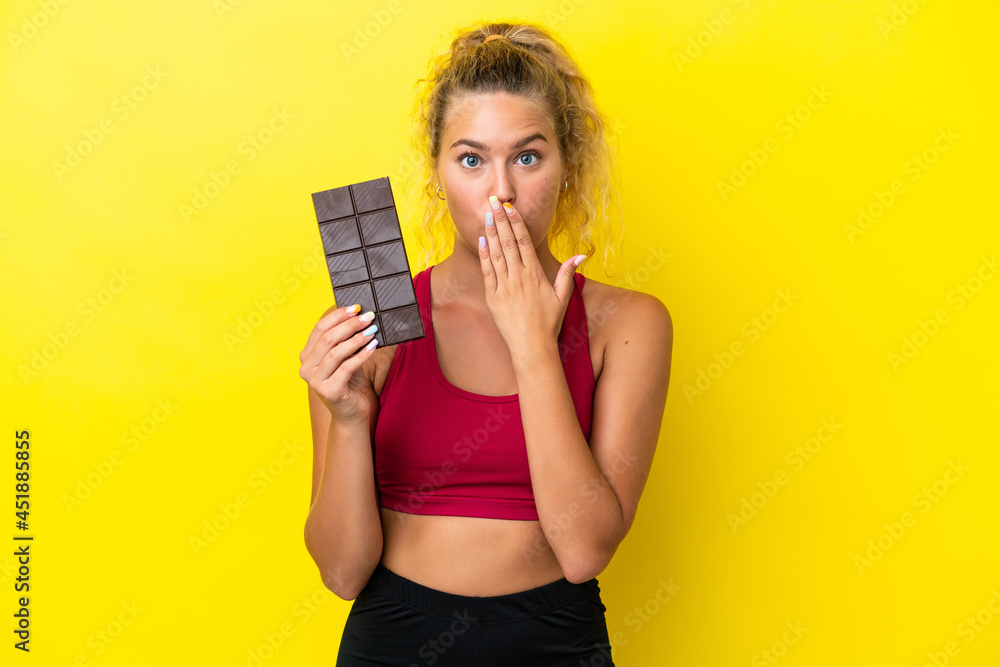 Girl with curly hair isolated on yellow background taking a chocolate tablet and surprised Girl with curly hair isolated on yellow background taking a chocolate tablet and surprised