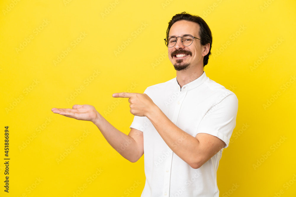 Young caucasian man isolated on yellow background holding copyspace imaginary on the palm to insert an ad Young caucasian man isolated on yellow background holding copyspace imaginary on the palm to insert an ad