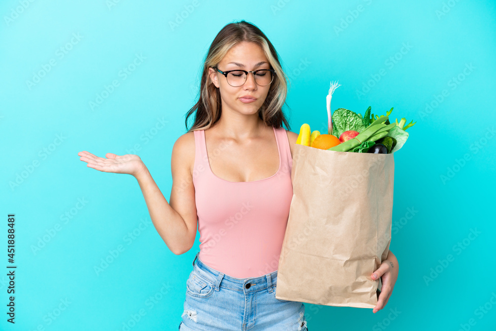 Young Russian woman holding a grocery shopping bag isolated on blue background having doubts while raising hands Young Russian woman holding a grocery shopping bag isolated on blue background having doubts while raising hands