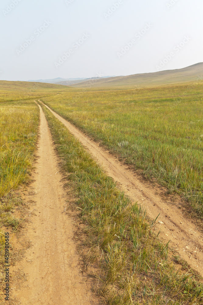 Dirt road on sandy soil in the steppe with green meadows on Olkhon island, Siberian Baikal Lake. Summer landscape. Travel and long distance path concept