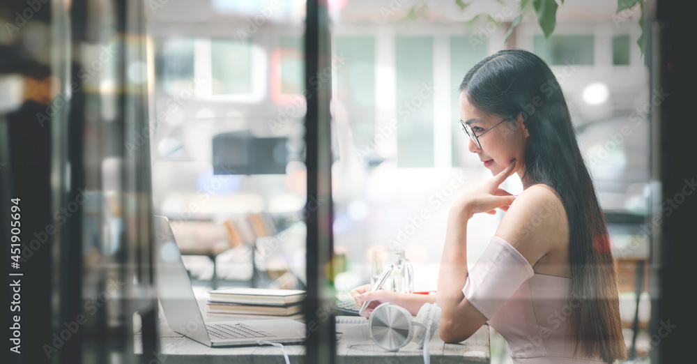 Side view of beautiful asian woman with glasses and long hair working at office desk, looking through window glass.