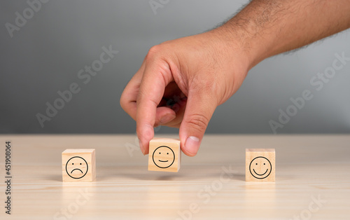 Hand picking one from three wooden blocks with emoticons, on wood table with gray background. Customer experience, satisfaction survey, service rating concept.