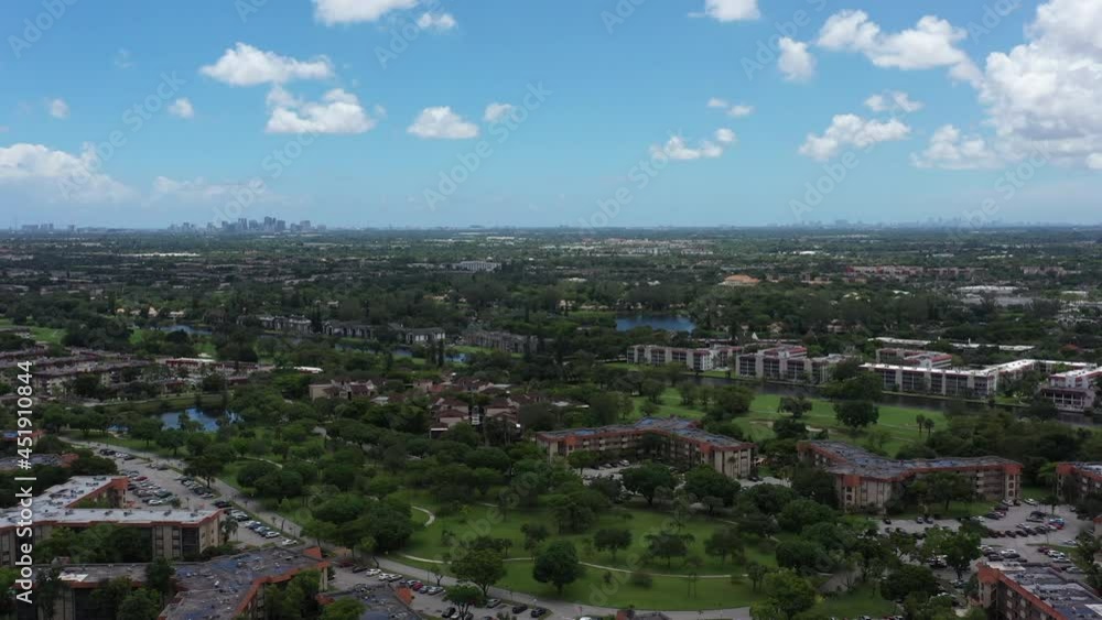 An aerial view over green trees and grass on a sunny day near apartments. The camera dolly in and boom down towards the trees with clouds above and more apartment buildings in the distance. An aerial view over green trees and grass on a sunny day near apartments. The camera dolly in and boom down towards the trees with clouds above and more apartment buildings in the distance.