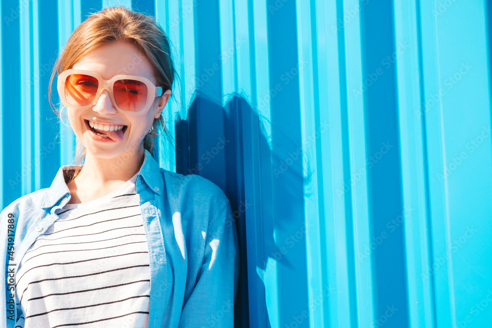 Young beautiful smiling hipster woman in trendy summer clothes. Sexy carefree woman posing on the street background near blue wall at sunset. Positive model outdoors. Cheerful and happy in sunglasses - obrazy, fototapety, plakaty 