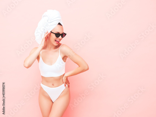 Young beautiful smiling woman in white lingerie. Sexy carefree model in underwear and towel on head posing pink wall in studio. Positive and happy female enjoying morning in sunglasses