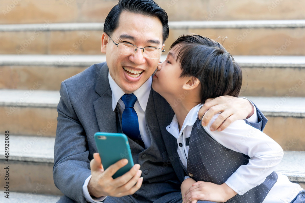 A business man wearing a suit. He was looking at the mobile phone in his hand and smiled happily while his lovely son was kissing his cheek - Dad and Son happy Asian family concept