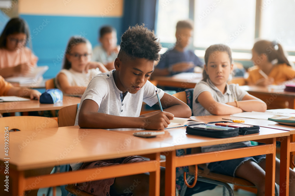 African American schoolboy writes while having class with his classmates at elementary school. African American schoolboy writes while having class with his classmates at elementary school.