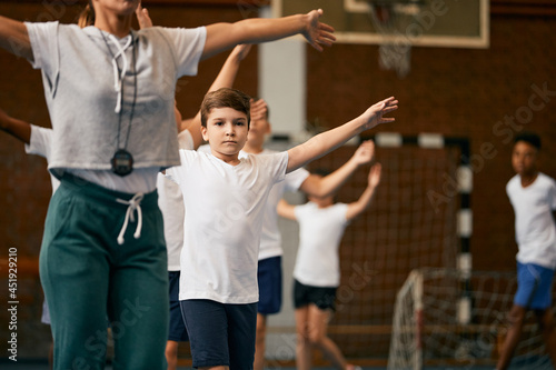 Elementary student and his friends doing warm-up exercises during PE class with their coach at school gym.