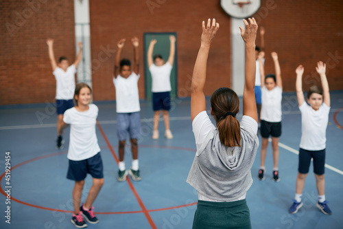 Rear view of coach and her students warming up during physical education at elementary school gym.