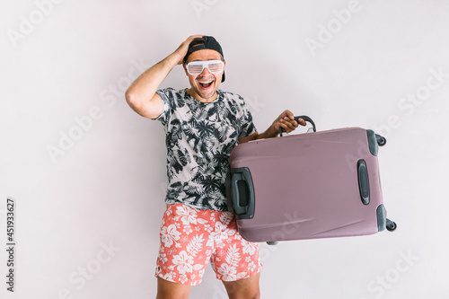 Young man in a floral shirt and cap, with a suitcase to go on vacation, in daylight on a white wall photo