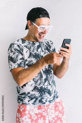 Young man in a floral shirt, cap and glasses in summer looking at his smartphone and drinking a cocktail with a straw, in daylight on a white wall photo