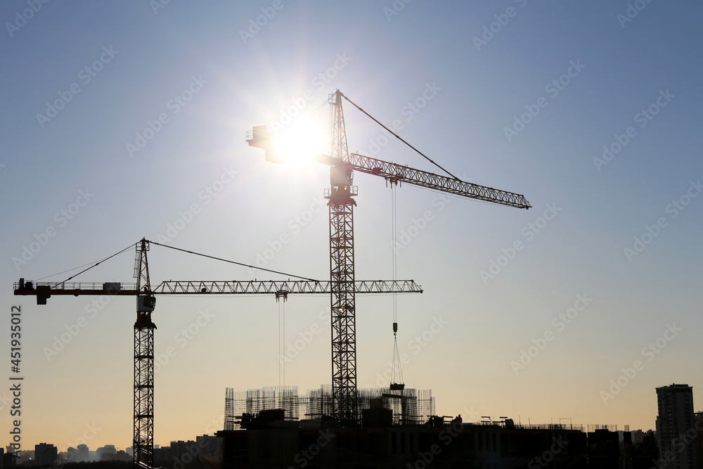 Silhouettes of construction cranes on unfinished residential building against the sky and shining sun. Housing construction, apartment block in city
