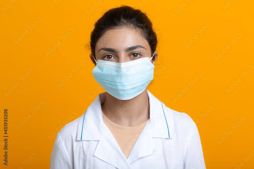 Portrait of indian doctor woman wearing white medical gown and protective surgical face mask isolated on yellow background. Portrait of indian doctor woman wearing white medical gown and protective surgical face mask isolated on yellow background.