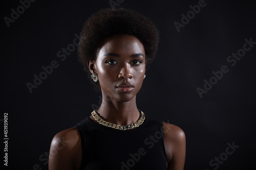 Portrait of young serious stylish black woman. Beautiful curly dark haired african-american girl wear black dress with gold jewelry and looking at camera. Studio shoot isolated on dark background.