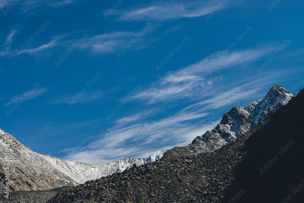 Awesome alpine landscape with dark rockies and high snow-covered mountains in sunshine under blue sky with cirrus clouds. Beautiful highland scenery with sharp black rocks and mountain top with snow.